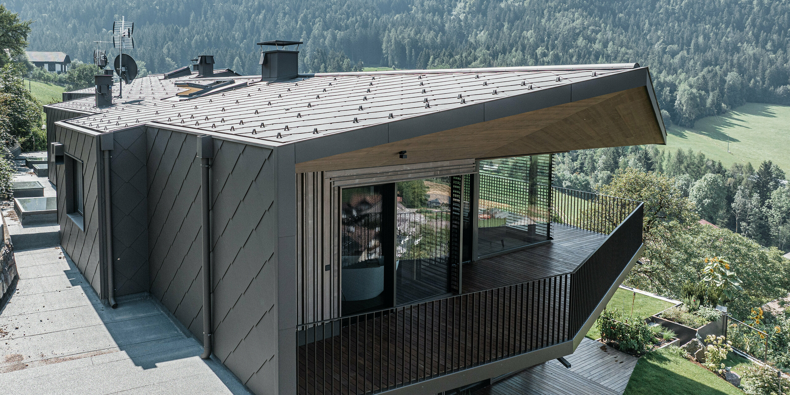 Doppelhaushälfte in Mölten (Südtirol, Italien) mit einer Kombination aus dunklen Aluminiumrauten und Holzverkleidung. PREFA Dach- und Wandrauten 44 x 44 in P.10 Braun bieten effektiven und ästhetisch ansprechenden Schutz. Das Farbe der Metallverkleidung geht nahtlos in die bewaldete Umgebung über. Der architektonische Entwurf betont eine offene Terrasse, die einen entspannenden Außenbereich schafft. Großzügige Glasfronten bieten einen einladenden Blick auf das schöne Bergpanorama. Das Schneeschutzsystem auf dem Dach verhindert in den Wintermonaten unkontrollierte Dachlawinen.