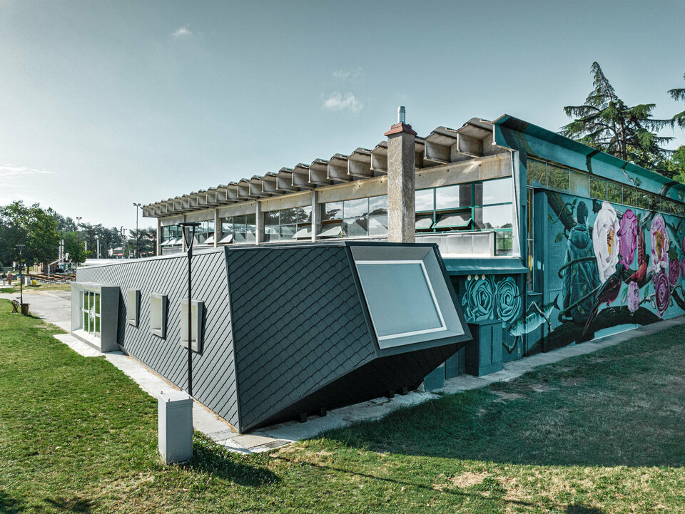 Die Turnhalle Rodriguez und ihr neuer Zubau mit der Rautenschuppenhaut aus PREFA Aluminium von der Seite; der Rasen erstreckt sich davor und zur Seite, im Hintergrund ist ein Teil eines Sportplatzes zu sehen.