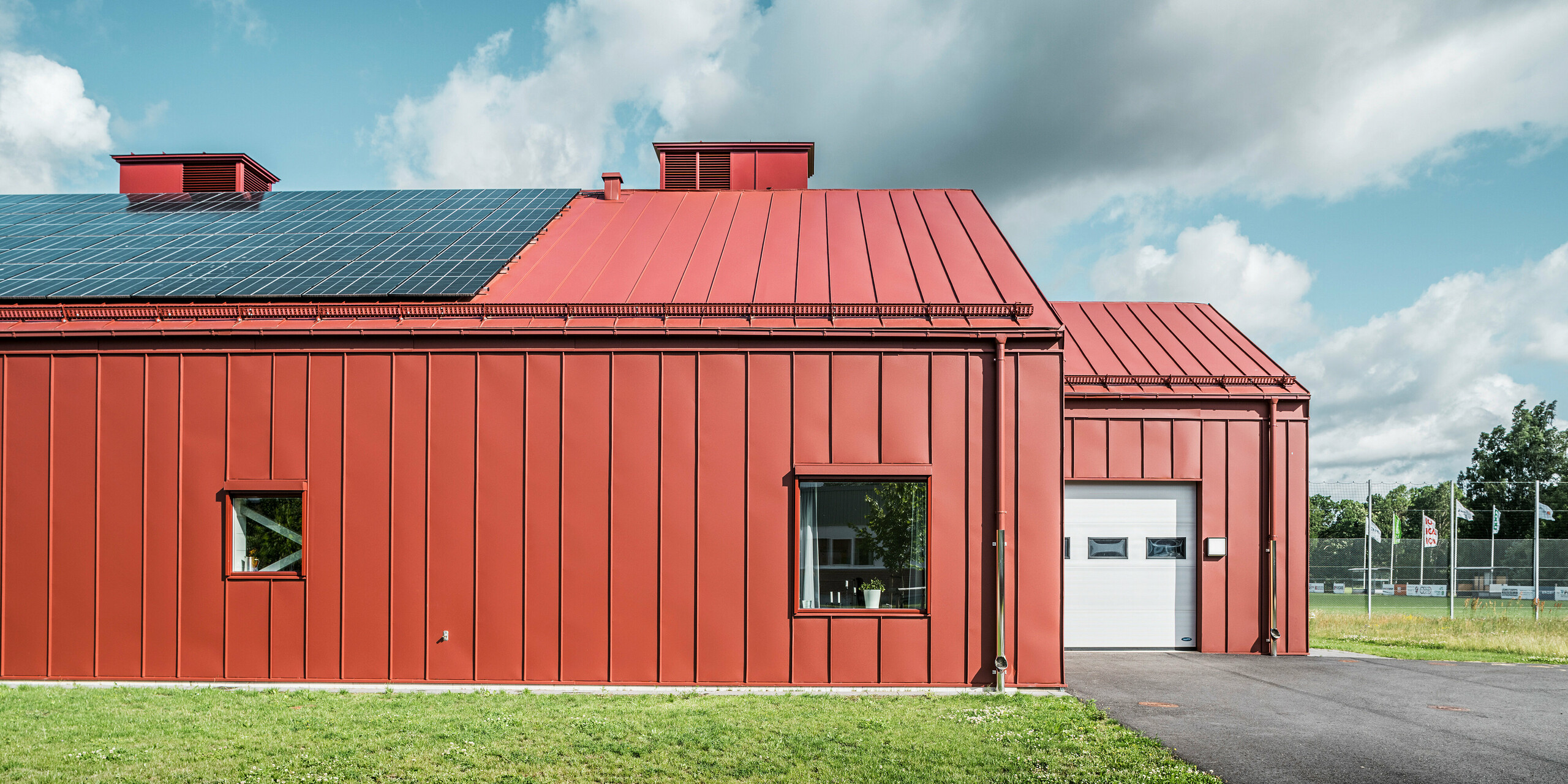 Seitenansicht des Restaurants Gröna Hjärtat, der Schulkantine der Göingeskolan und Prästavångskolan in Broby, Schweden. Die Perspektive zeigt die markante Fassade und das Dach mit Stehfalzdeckung aus PREFALZ in Oxydrot. Eine großflächige Photovoltaikanlage auf dem Dach erzeugt Strom aus erneuerbare Energie. Die langlebige Aluminiumverkleidung in Oxydrot unterstreicht das moderne und energieeffiziente Design des Bauwerks. Die PREFA Dachentwässerung ist harmonisch in die Fassade integriert, passend zur gesamten Gebäudeoptik.