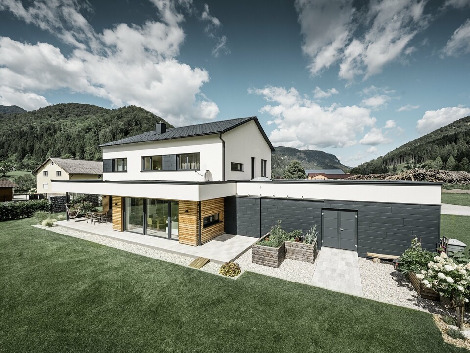 Das Bild zeigt ein modernes Einfamilienhaus in einer idyllischen Landschaft mit einer klaren Sicht auf bewaldete Hügel im Hintergrund. Das Haus zeichnet sich durch eine Kombination aus weißen Putzwänden und einer markanten Fassadenverkleidung mit PREFA Siding.X in P.10 Anthrazit aus, die für einen modernen und langlebigen Look sorgt. Die dunkle Färbung der Fassadenelemente bildet einen ansprechenden Kontrast zum hellen Außendesign und zur natürlichen Umgebung. Die Anordnung der Siding.X Fassadenpaneele ist präzise und verleiht der Immobilie einen strukturierten und hochwertigen Charakter, der typisch für PREFA Produkte ist. Das Dach des Gebäudes wurde mit der bewährten PREFA Dachplatte eingedeckt. Die Dachentwässerung erfolgt durch das Ablaufrohr und die Hängerinne von PREFA.