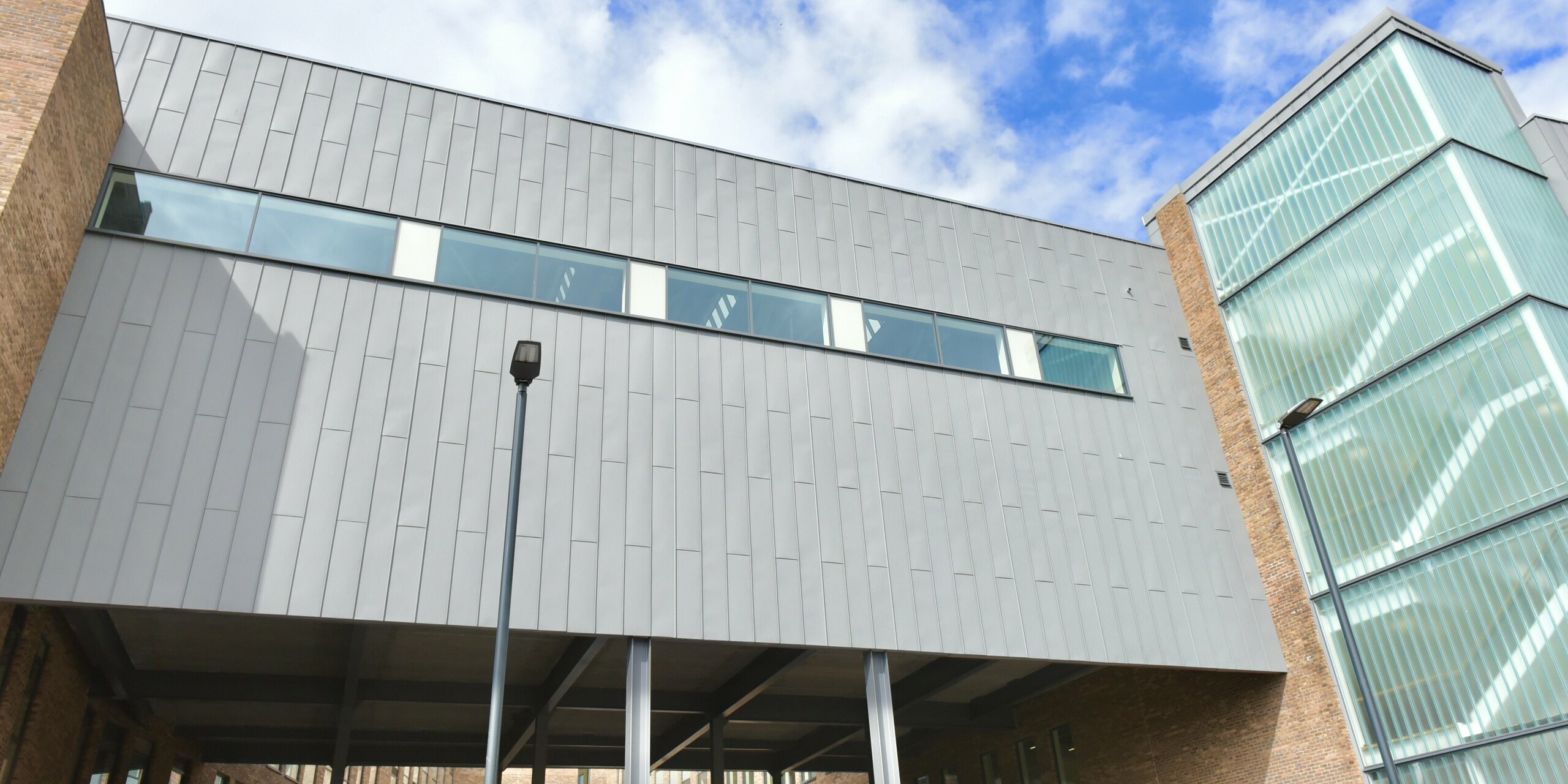 Gebäudebrücke des North Regional College in Coleraine mit PREFALZ Aluminiumfassade in Patinagrau. Die innovative Stehfalzdeckung verleiht der Bildungseinrichtung Leichtigkeit und zugleich Robustheit. Die Fassade aus Aluminium und Ziegel harmoniert und verleiht dem Gebäude eine zeitlose Architektur.