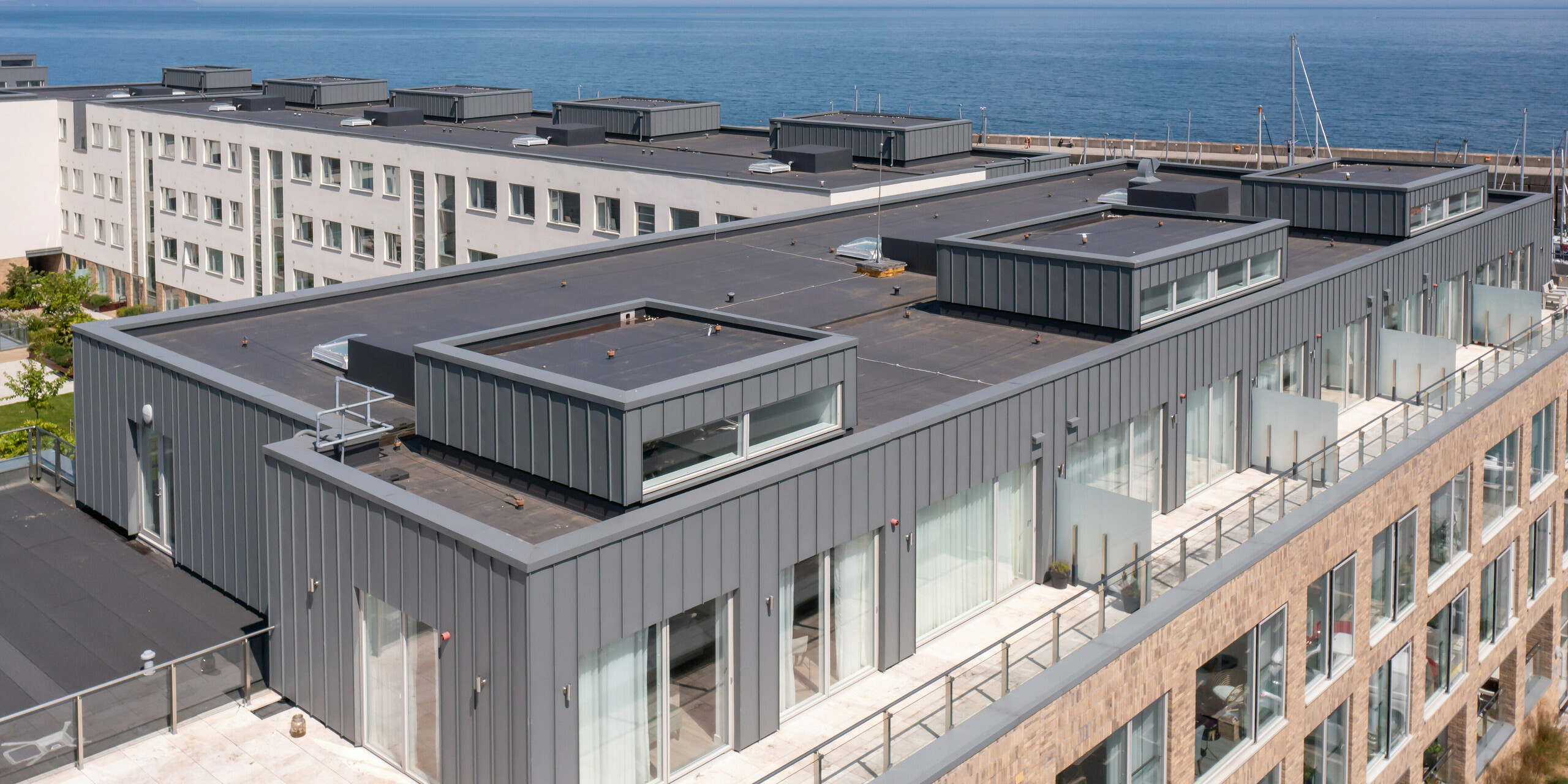 Marina Village Greystones in Wicklow, Irland – Vogelperspektive auf die oberste Etage von einem der modernen Wohngebäude. Das Bild zeigt die detaillierte Ansicht der unbewohnten Dachterrassen. Im Hintergrund ist das strahlend blaue Wasser des Yachthafens zu erkennen. Die Verkleidung des Stockwerks besteht aus hochwertigem PREFALZ in der Farbe P.10 Hellgrau. Die robuste Aluminiumverkleidung bietet dauerhaften Schutz und eine ästhetische Optik. Große Glasfenster bieten den zukünftigen Bewohnern einen atemberaubenden Blick auf das Meer und den Yachthafen. Die Perspektive zeigt bewusst die beeindruckende Architektur in der malerischen Kulisse des Yachthafens.