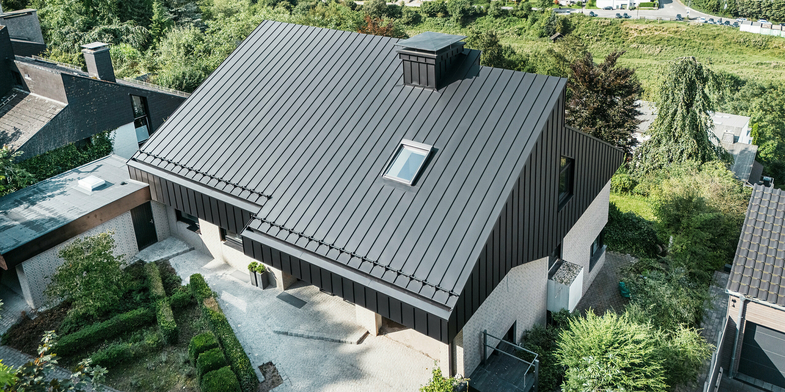 Blick von oben auf ein modernes Einfamilienhaus in Meschede, Deutschland, mit einem versetzten Pultdach aus PREFALZ Aluminium in P.10 Schwarz. Das Aluminiumdach und die elegante Aluminiumfassade an den Giebeln sind präzise verarbeitet und unterstreichen so die ästhetische und witterungsbeständige Gestaltung. Im Hintergrund erstreckt sich eine idyllische ländliche Umgebung mit grünen Hügeln und einer ruhigen Wohnsiedlung.