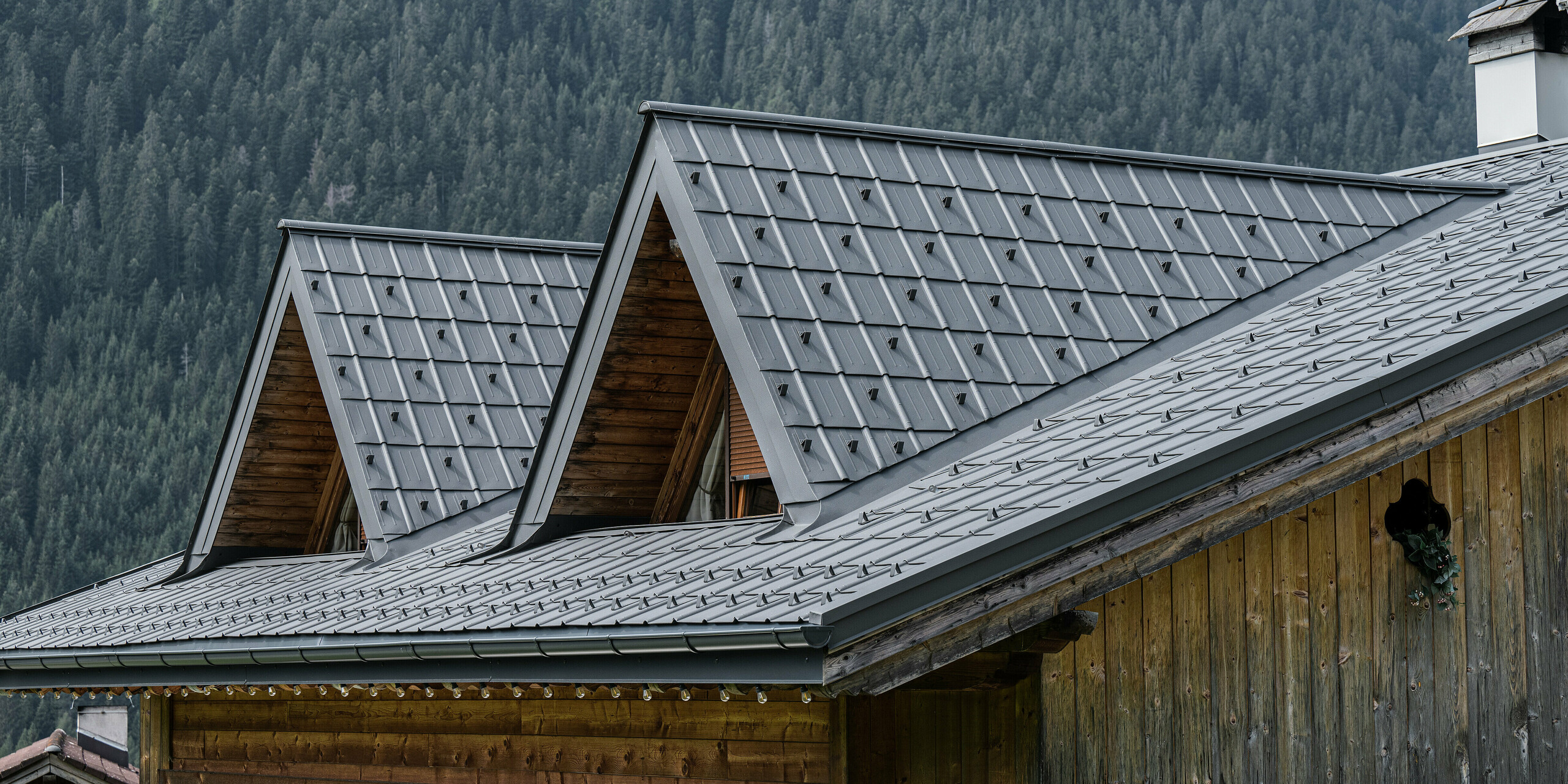 Nahaufnahme des Alpenhauses in Auronzo di Cadore mit dem neu verlegten PREFA Dach in P.10 hellgrau. Die Dachplatten aus hochwertigem Blech verleihen dem traditionellen alpinen Baustil einen modernen Touch. Mit ihrer Textur und Farbe schaffen die Aluminiumprodukte eine harmonische Verbindung mit der umgebenden Waldlandschaft. Dieses Bild veranschaulicht die Kombination von regionaler Handwerkskunst mit innovativen und langlebigen Baumaterialien.