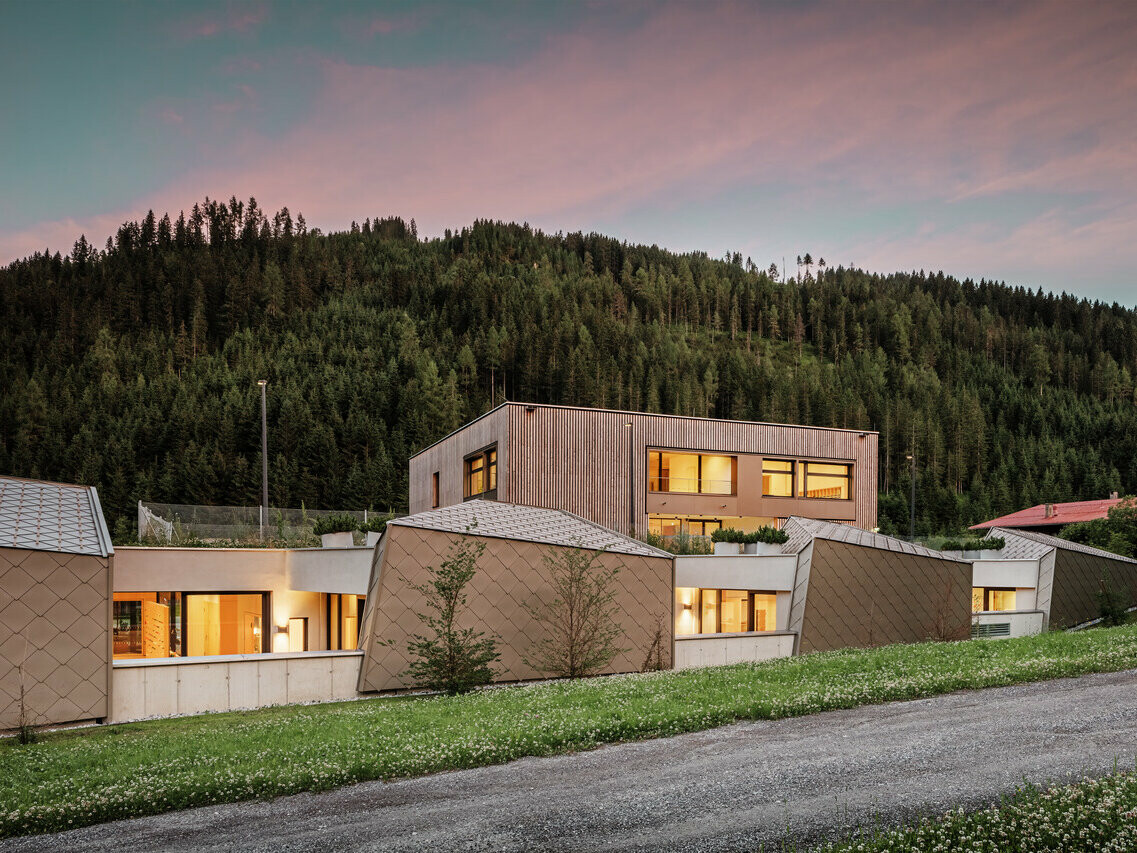 Das Bild zeigt das Gerlos-Bildungszentrum in der Abenddämmerung, mit Innenbeleuchtung. Mit seiner modernen Holzverkleidung und den rautenförmigen Metallpaneelen fügt sich das Gebäude harmonisch in die natürliche Umgebung des dichten Nadelwaldes im Hintergrund ein. Der Himmel ist in sanfte Abendtöne getaucht und schafft eine ruhige und einladende Atmosphäre.