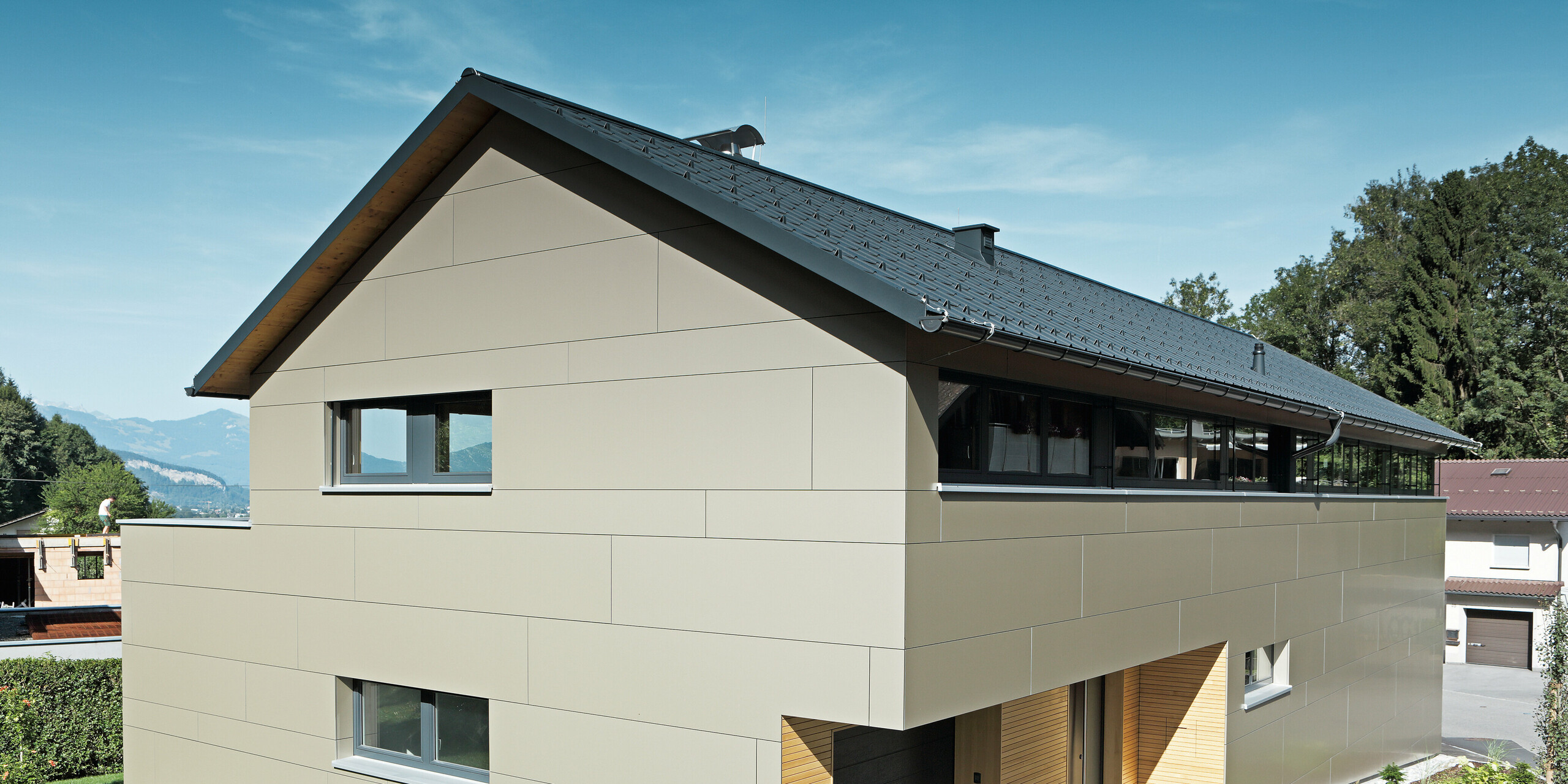 Elegantes Einfamilienhaus in Hohenems mit PREFA Komplettsystem: Die Fassade aus Aluminium Verbundplatten in Bronzemetallic und das Dach aus PREFA Dachplatten in P.10 Anthrazit fügen sich nahtlos in die umgebende Landschaft ein. Die klare, strukturierte Architektur mit dem sichtbaren Holzunterdach, kombiniert mit der robusten, wartungsarmen Aluminiumverkleidung, bietet einen nachhaltigen und ästhetischen Wohnraum, eingebettet in die natürliche Umgebung mit Bergblick im Hintergrund.