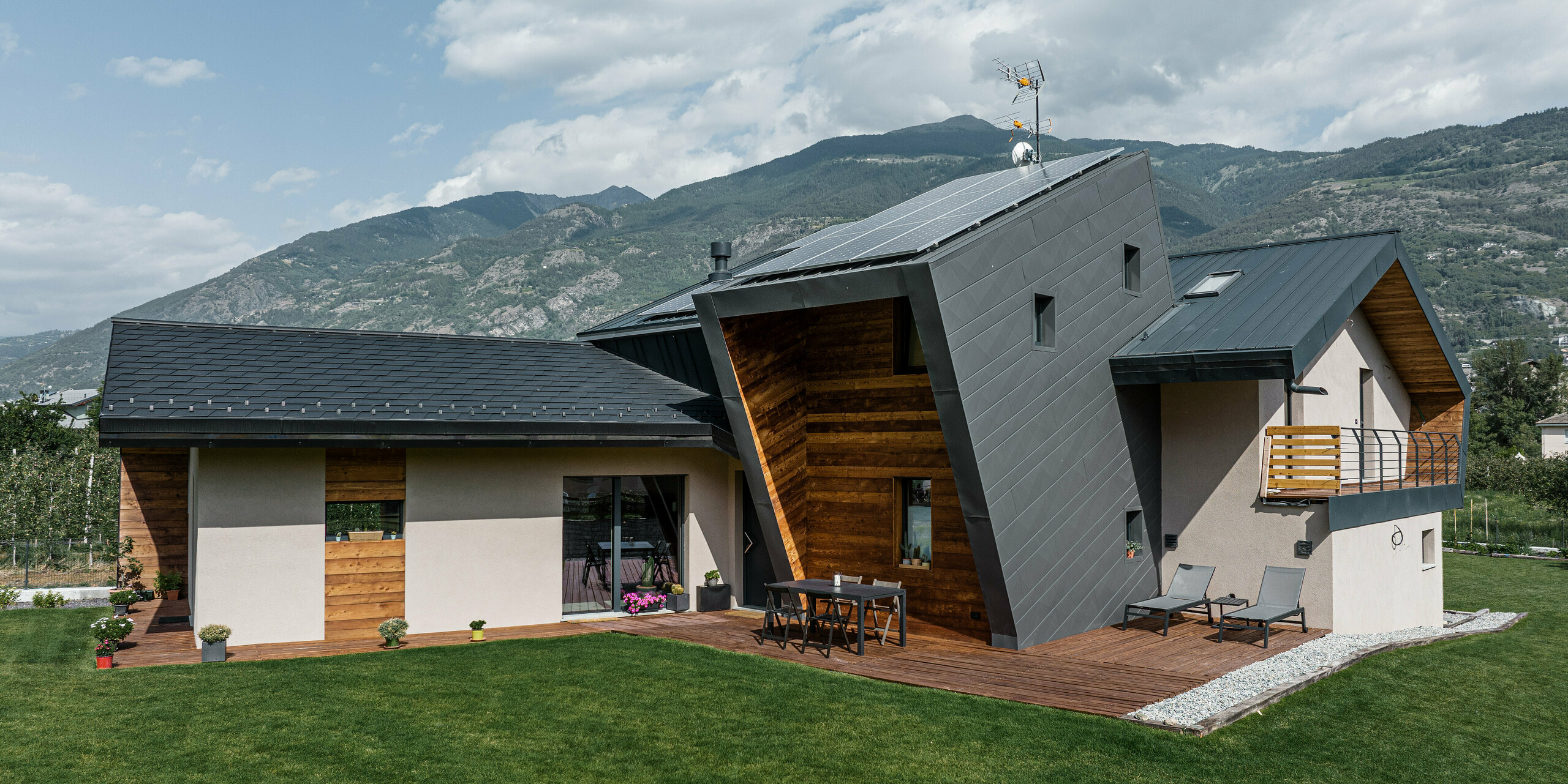 Die Villa Tia in Italien, architektonisch bereichert mit einem PREFA Komplettsystem, präsentiert sich mit einer eleganten Kombination aus dunklen PREFA Aluminiumprodukten. Das Satteldach und Teile der Fassade sind mit PREFALZ in P.10 Anthrazit eingedeckt. Das Nebengebäude hebt sich mit schwarzen Dachschindeln ab. Die Verbindung aus der hellen Putzfassade und natürlichen Holzelementen unterstreicht die moderne Eleganz. Ein besonderer Blickfang ist der Bereich der Terrasse, wo als spezielle Lösung schräg liegende Siding.X Fassadenpaneele in P.10 Dunkelgrau das Design vervollständigen. Die Aluminiumelemente garantieren dabei nicht nur eine optische Aufwertung, sondern versprechen auch Langlebigkeit und Nachhaltigkeit.