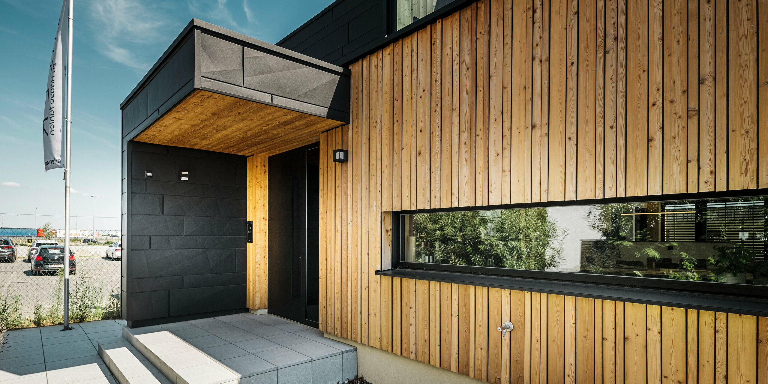 Seitlicher Eingang des Musterhauses Autark im Fertighauszentrum Blaue Lagune in Vösendorf, mit einer modernen Fassade aus natürlichem Holz und schwarzen PREFA Siding.X Paneelen. Das Bild zeigt die edle Kombination aus Holzverkleidung und Aluminium in der hochwertigen Beschichtung P.10 Schwarz.