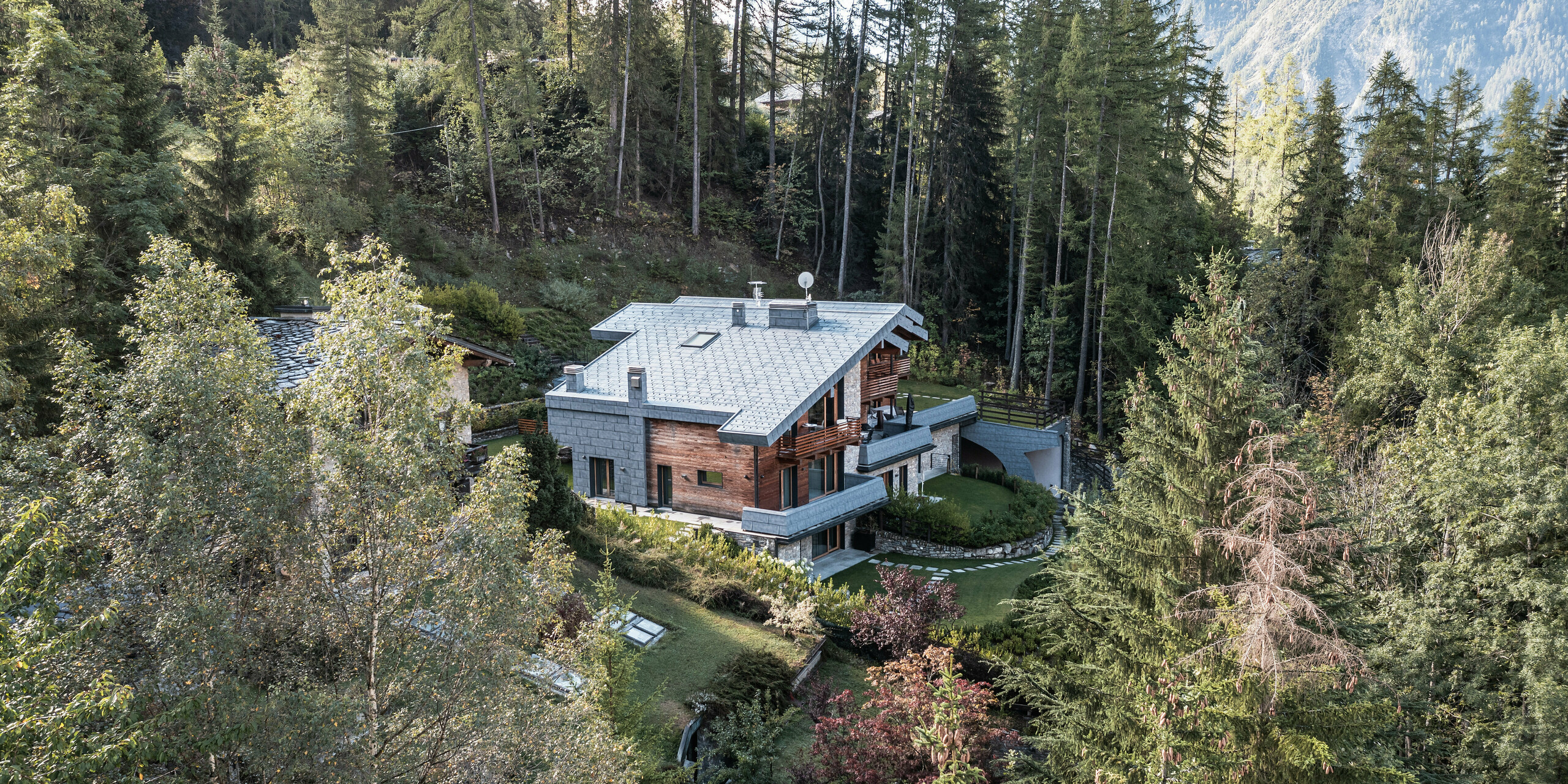 Eine Luftaufnahme einer beeindruckenden Villa in Courmayeur, umgeben von dichten Wäldern, die die natürliche Architektur aus Holz, Stein und PREFA Aluminium hervorhebt. Die schieferfarbenen Dach- und Fassadenpaneele FX.12 von PREFA dienen als Verbindungselement und schützen das Gebäude langfristig. Die abgestufte Anordnung der Gebäudeteile ermöglicht die Integration in die steile Landschaft, während mehrere Balkone und gepflegte Gärten einen angenehmen Außenbereich bieten, der zum Entspannen in der ruhigen alpinen Umgebung einlädt.