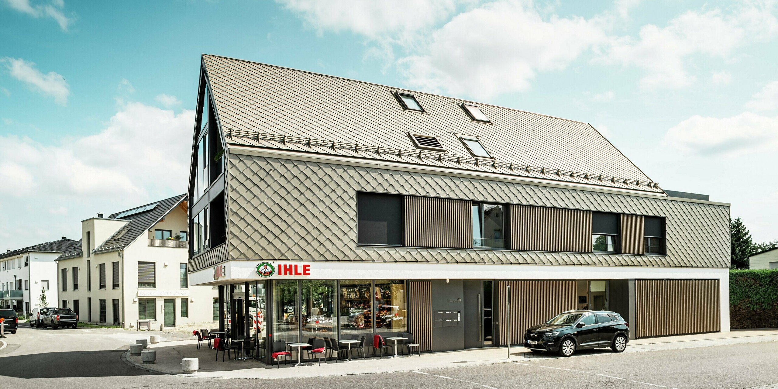 Mehrfamilienhaus in Leitershofen, Deutschland, mit integrierten Gastronomieflächen im Erdgeschoss. Das architektonisch anspruchsvolle Bauwerk wurde mit stilvollen PREFA Dach- und Wandrauten 29 × 29 in Bronze eingedeckt. Die Aluminiumrauten verleihen dem Gebäude eine elegante und witterungsbeständige Hülle. Große Glasflächen und Holzelemente unterstreichen das durchdachte Design, während die weiße Putzfassade im Erdgeschoss einen klaren Kontrast bildet. 