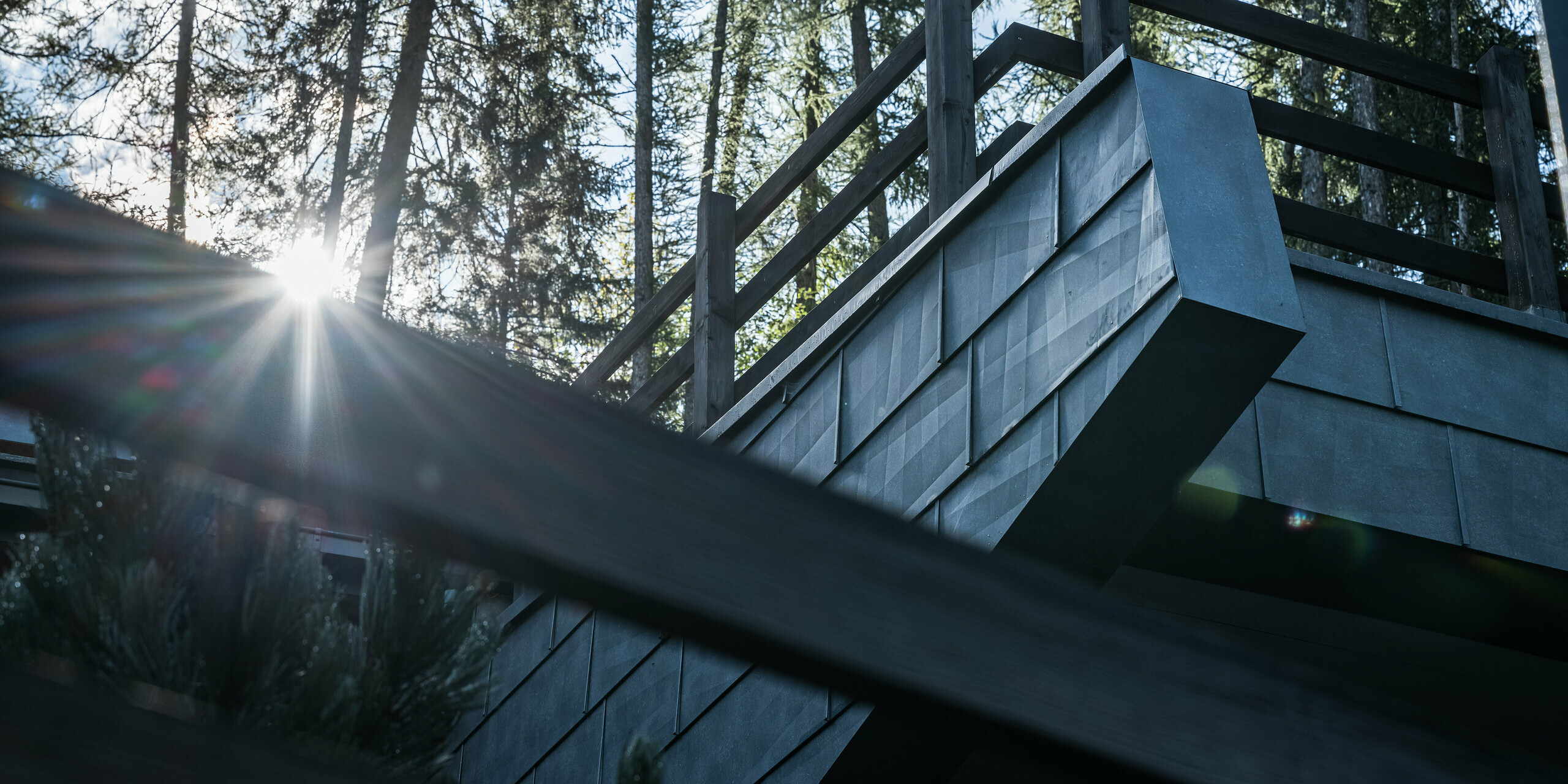 Nahaufnahme des Holzgeländers und der eleganten PREFA FX.12 Fassadenelemente aus Aluminium. Die Fassadenpaneele in P.10 Steingrau sind an einer Villa in Courmayeur, Italien, installiert. Die Sonne scheint durch die Kiefern und erzeugt ein schillerndes Lichtspiel, das die strukturierte Oberfläche der Aluminiumfassade hervorhebt und dramatische Schatten wirft, die die robuste und ästhetische Struktur betont.