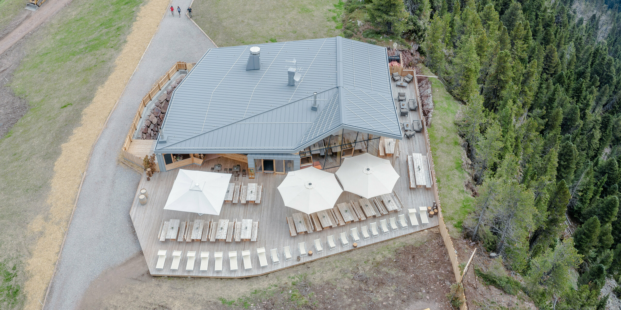 Luftaufnahme des Ristorante Busabelle, umgeben von sanften Wanderwegen und dichtem Wald. Die Architektur des Restaurants zeigt eine Kombination aus moderner hellgrauer PREFALZ Metallfassade und natürlichem Holz, die in die alpine Landschaft eingebettet ist. Die großflächige Terrasse ist ausgestattet mit Holzbänken und großen Sonnenschirmen, die einen komfortablen Aufenthaltsbereich für Gäste in dieser idyllischen Bergumgebung schaffen. Das Gebäude mit der Hülle aus Blech ist besonders robust und langlebig.