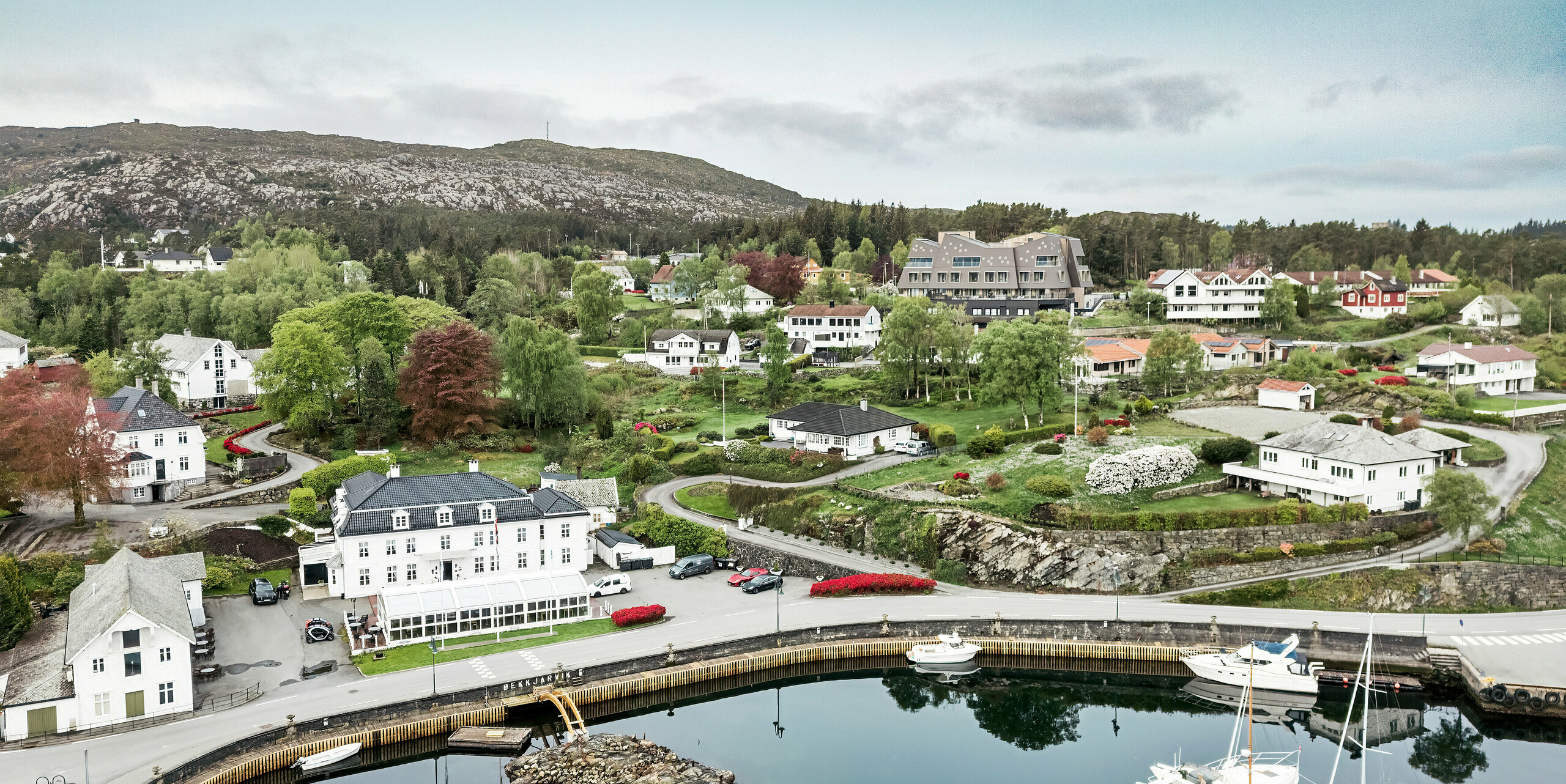 Luftaufnahme des Hafens von Bekkjarvik, Norwegen, mit Blick auf das Beckerwyc House im Hintergrund. Das Hotel hebt sich durch seine futuristische Architektur und die Fassade aus PREFA Wandrauten 44 × 44 in Nussbraun von den umliegenden traditionellen weißen Holzhäusern ab. Die malerische Landschaft, der Hafen mit den Booten und die grüne Umgebung unterstreichen die einzigartige Lage des Hotels in dieser idyllischen norwegischen Küstenstadt auf der Insel Selbjørn.