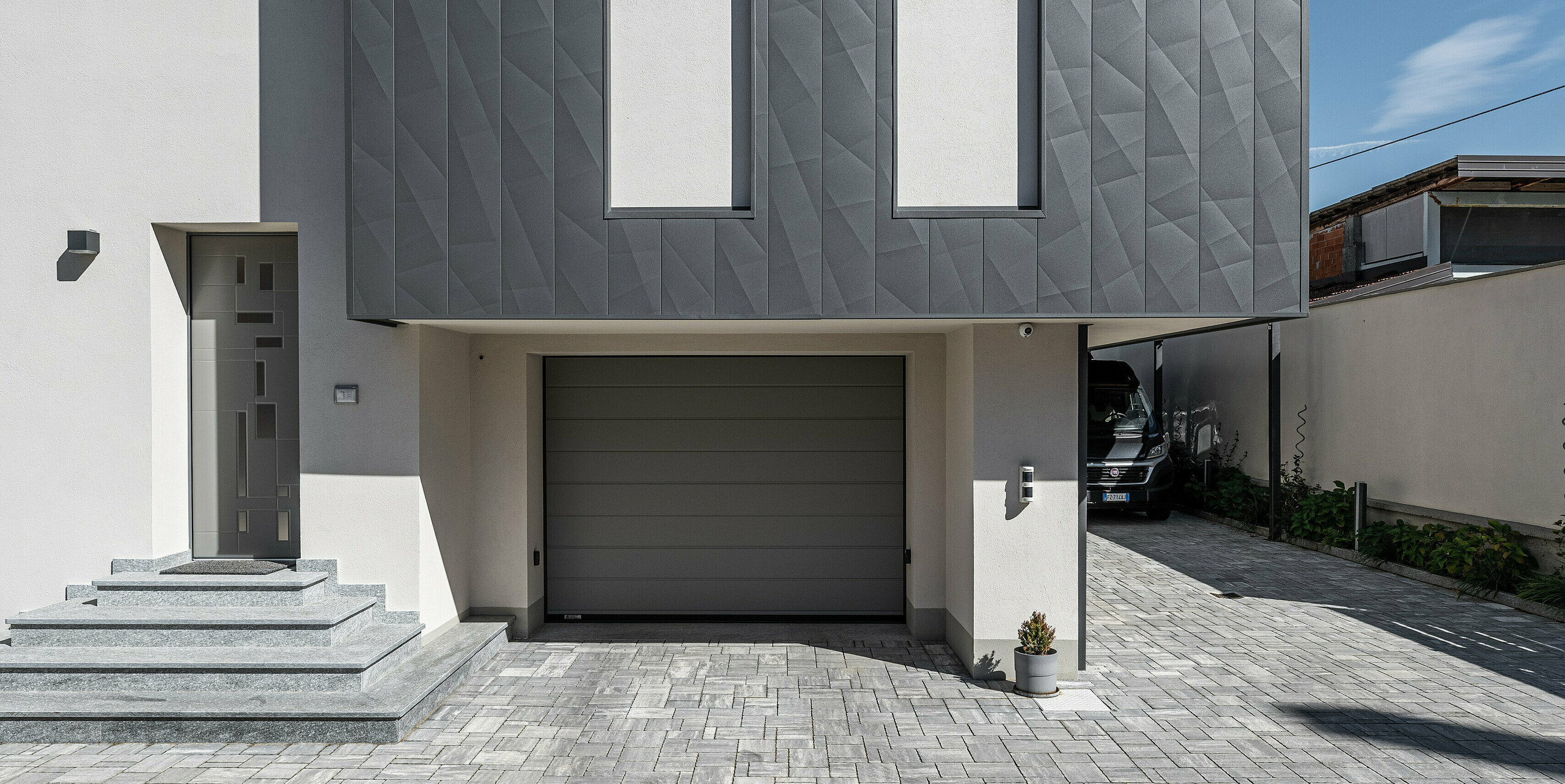 Frontansicht der Garage eines modernen Einfamilienhauses in Savigliano, Italien, hervorgehoben durch eine PREFA Siding.X Aluminiumfassade in P.10 Dunkelgrau. Die geometrische Textur der Fassade verleiht dem Gebäude ein markantes und zeitgemäßes Aussehen, während die rechteckigen Fenster und die graue Garagentür eine klare und funktionale Ästhetik unterstützen. Die sauber gepflasterte Einfahrt rundet das elegante Erscheinungsbild ab und sorgt für einen nahtlosen Übergang vom öffentlichen zum privaten Raum.