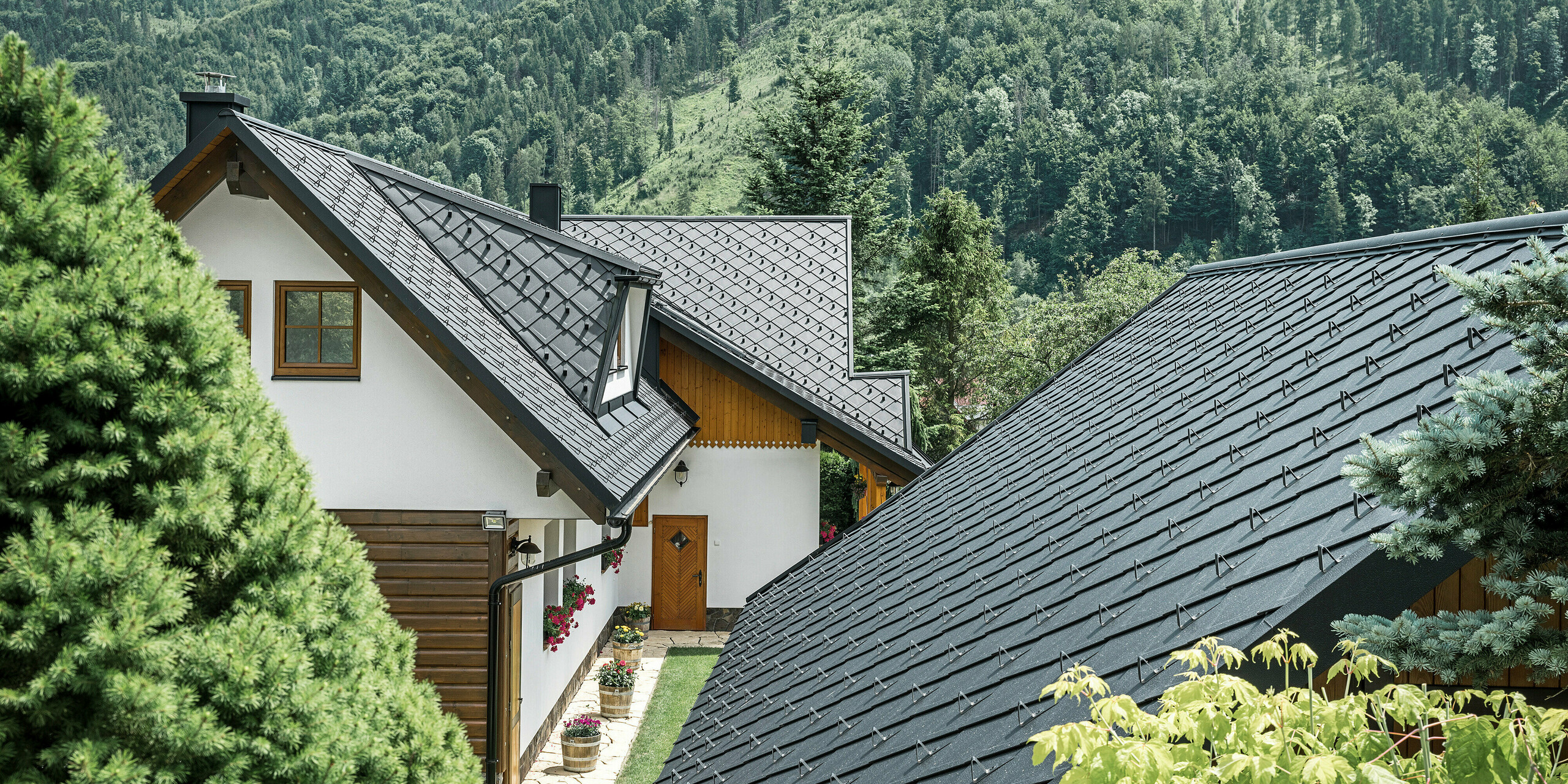 Rückansicht eines traditionellen Einfamilienhauses in Stara Bystrica, Slowakei, mit einer atemberaubenden Dachlandschaft aus PREFA Dachrauten 29 × 29 in P.10 Schwarz. Die robusten Aluminiumdachziegel in Schuppenoptik harmonieren perfekt mit der natürlichen Umgebung und bieten einen modernen Kontrast zur bewaldeten Hügellandschaft im Hintergrund. Die detailliert ausgeführten Dachflächen und die gut integrierten Trapezgauben verleihen dem Gebäude eine einzigartige, ästhetische Anmutung.