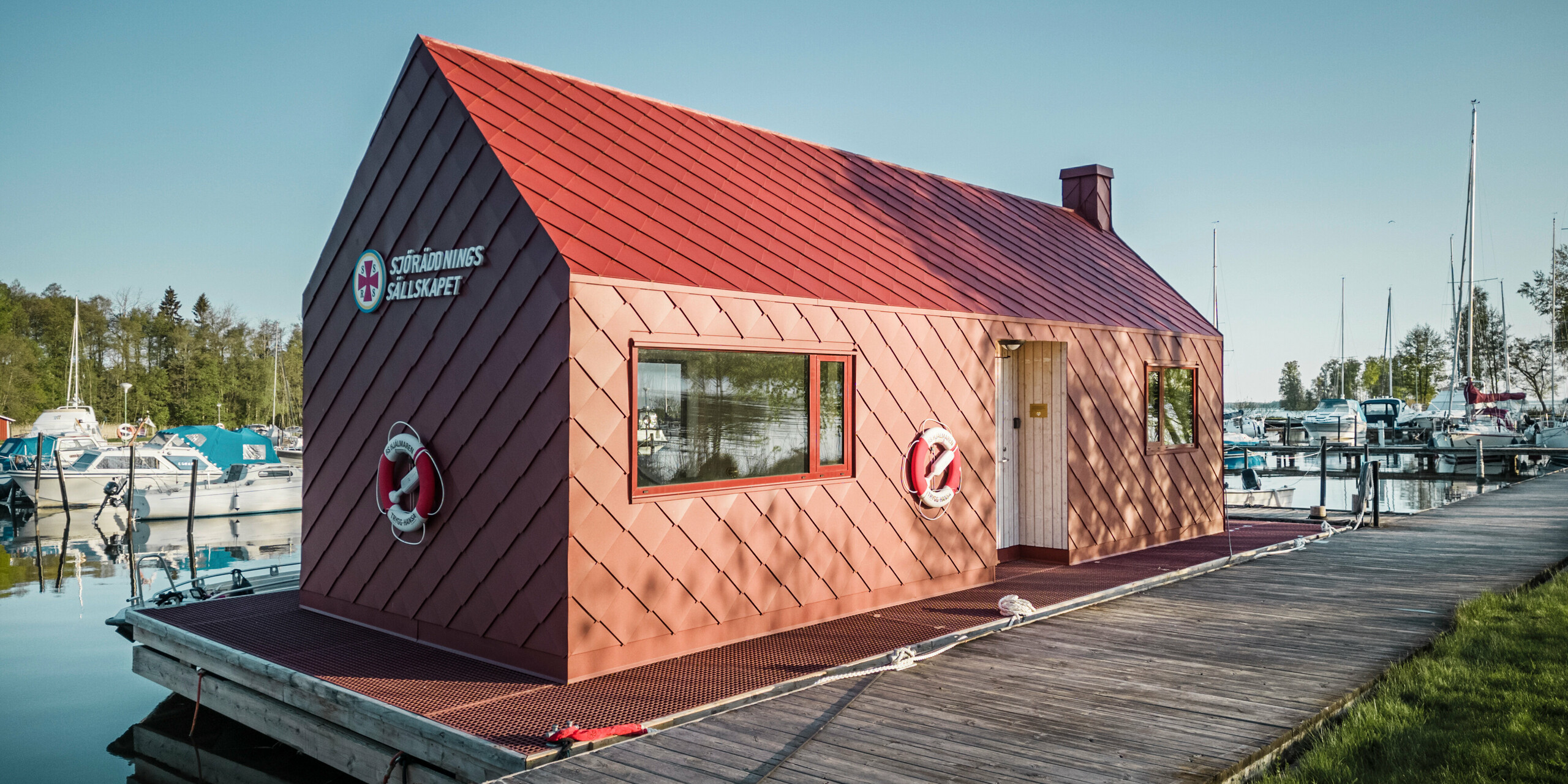 Die Seenotrettungsstation am Hjälmaren See in Schweden, festgemacht an einem Holzsteg. Das markante Gebäude mit einer Fassade aus oxydroten PREFA Dach- und Wandrauten 29×29 steht stolz im Kontrast zu den weißen und blauen Booten im Hintergrund. Mit Rettungsringen ausgestattet und bereit für den Einsatz, vermittelt das Haus sowohl Sicherheit als auch einen starken maritimen Charakter in dieser friedvollen Seenlandschaft.