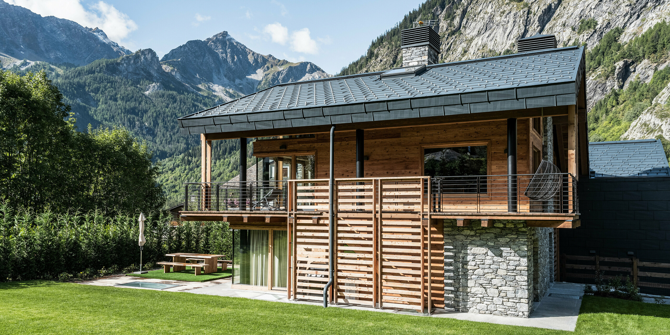 Luxusvilla in Courmayeur mit einer Fassade aus Naturstein und Holz und einem eleganten Dach aus Aluminiumplatten PREFA FX.12 in P.10 Steingrau. Die großzügigen Balkone bieten einen atemberaubenden Blick auf die umliegenden Berge. Der gepflegte Garten mit Sitzgelegenheit und sorgfältig gepflegtem Rasen lädt zum Verweilen in der beeindruckenden Alpenlandschaft ein.