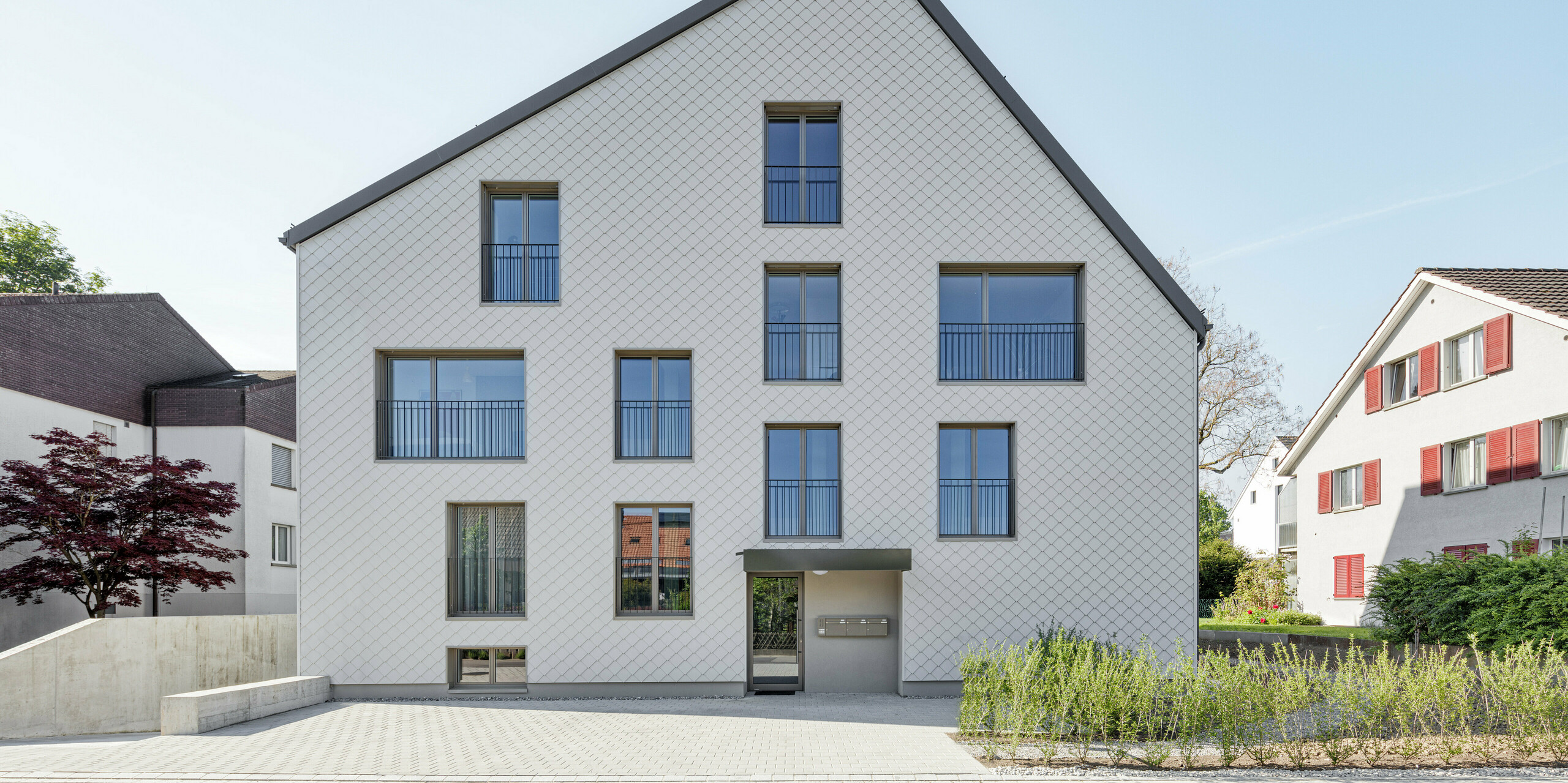 Blick auf den Eingangsbereich eines modernen Mehrfamilienhaus in Wiesendangen, Schweiz. Das Gebäude wurde mit einem hochwertigen PREFALZ Dachsystem in P.10 Dunkelgrau und einer eleganten Fassade aus PREFA Wandrauten in P.10 Prefaweiß verkleidet. Die saubere und geometrische Architektur spiegelt sich in der präzisen Anordnung der Wandrauten und der sorgfältigen Platzierung der Fenster wider, was zu einer attraktiven und zeitgenössischen Wohnästhetik führt. Das Haus ist harmonisch in seine Umgebung eingebettet, mit einem gepflegten Eingangsbereich und umgebenden Grünflächen, die die natürliche Schönheit und die nachhaltige Bauweise betonen.