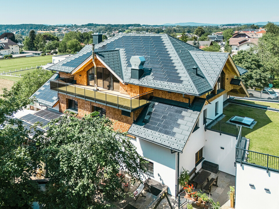 Vogelperspektive auf die Zenz'n Stub'n in Schörfling am Attersee mit neu saniertem Dach aus PREFA Solardachplatten und Dachplatten R.16 in P.10 Anthrazit. Das hochwertige Blechdach verbindet modernes Design mit nachhaltiger Energiegewinnung. Der großzügige Balkon und die harmonisch integrierte Dachentwässerung unterstreichen das stilvolle Erscheinungsbild des Gebäudes.