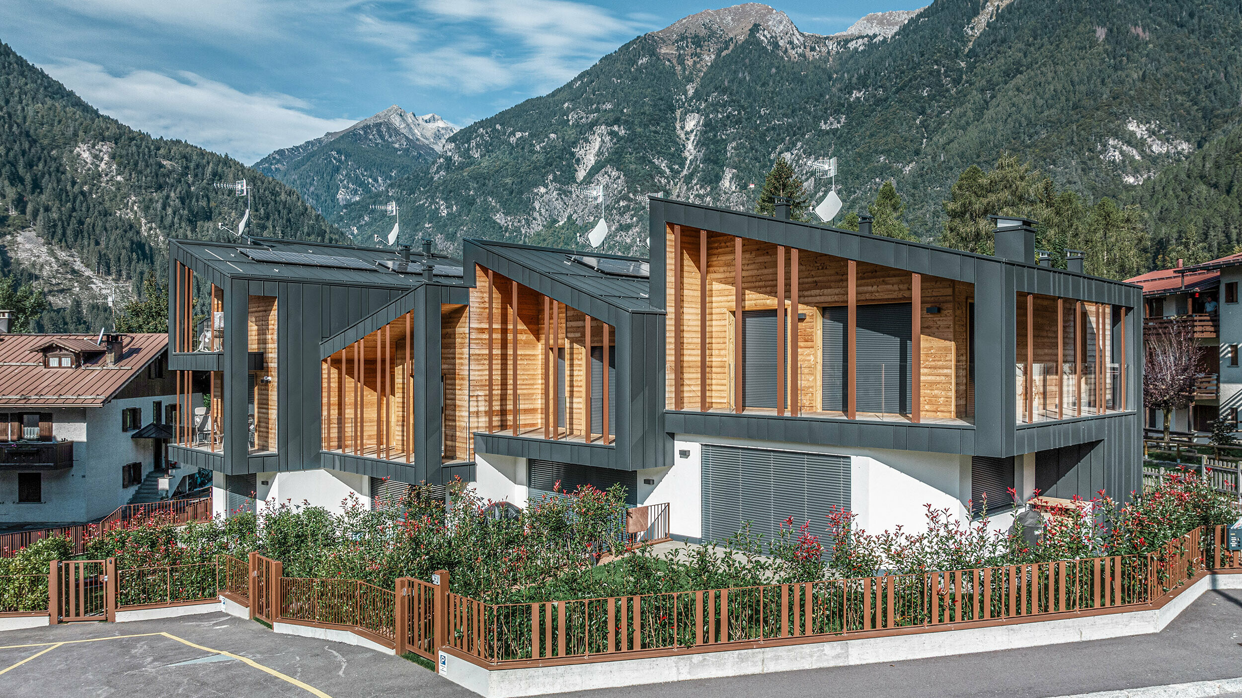 Neubau von modernen Chalets mit anthrazitfarbenen Prefalz-Metalldächern im Pultdachstil, Fassaden aus einer Kombination aus dunkelgrauem Metall und Holzelementen, in einer bergigen Landschaft mit klarem Himmel. Die großzügigen Fenster und Terrassen werden von grüner Bepflanzung und einem umzäunten Parkplatz ergänzt.