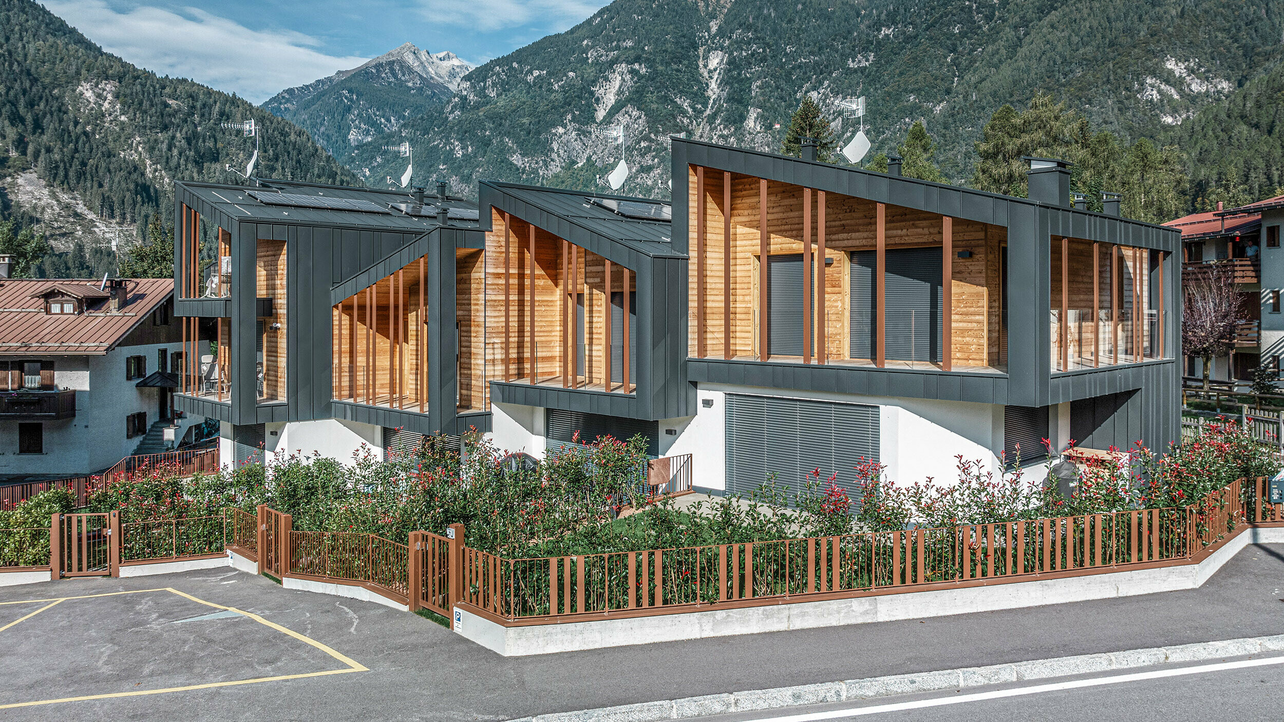 Neubau von modernen Chalets mit anthrazitfarbenen Prefalz-Metalldächern im Pultdachstil, Fassaden aus einer Kombination aus dunkelgrauem Metall und Holzelementen, in einer bergigen Landschaft mit klarem Himmel. Die großzügigen Fenster und Terrassen werden von grüner Bepflanzung und einem umzäunten Parkplatz ergänzt.