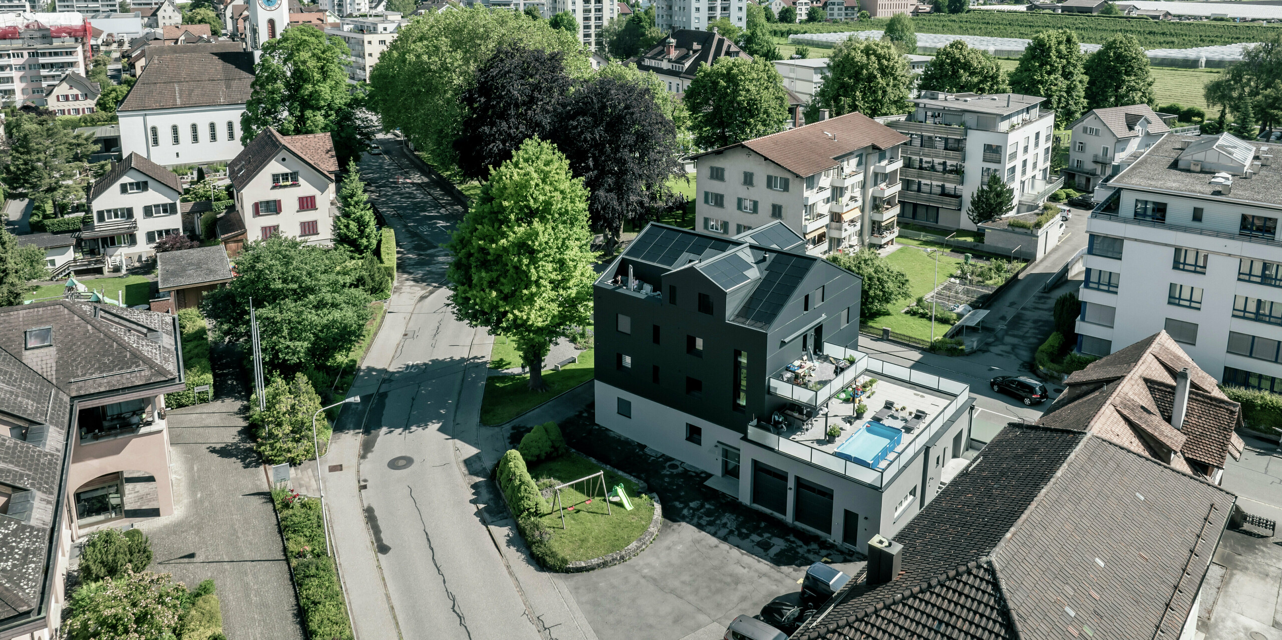 Vogelperspektive auf ein modernes Geschäftsgebäude mit einer sanierten Fassade aus PREFABOND Aluminium-Verbundplatten in Anthrazit. Das Gebäude fügt sich harmonisch in die grüne Umgebung und die umliegenden Wohnhäuser ein, während im Hintergrund eine beeindruckende Berglandschaft zu sehen ist.