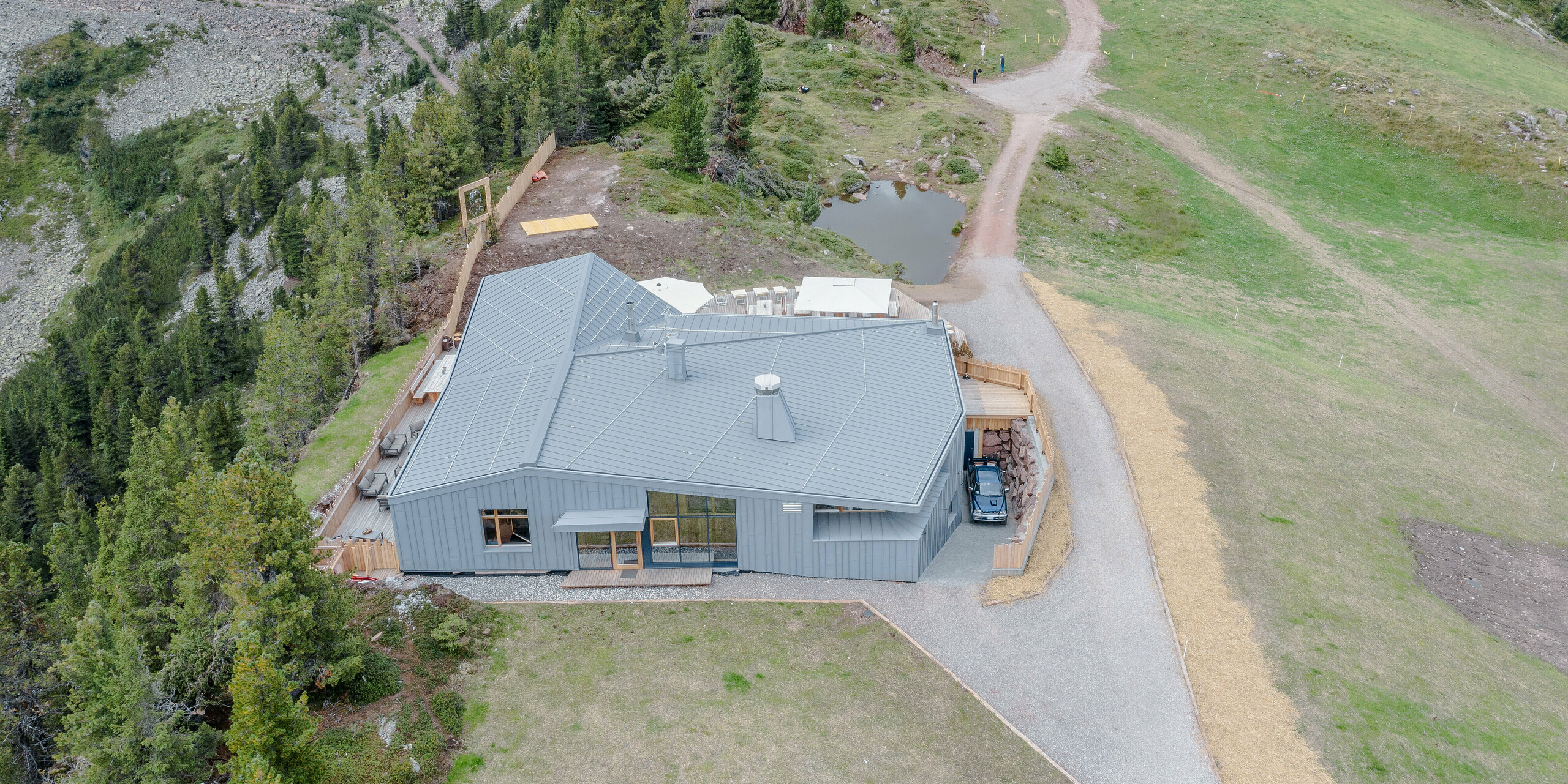 Vogelperspektive des Ristorante Busabelle, gelegen in einer majestätischen Berglandschaft. Das Restaurant ist durch sein modernes Design mit einer hellgrauen PREFALZ Dach- und Fassadenverkleidung aus Aluminium gekennzeichnet, die eine elegante und nahtlose Integration in die natürliche Umgebung ermöglicht. Wellige Wanderwege und ein kleiner Teich neben dem Restaurant betonen die malerische und ruhige Atmosphäre des Standorts.