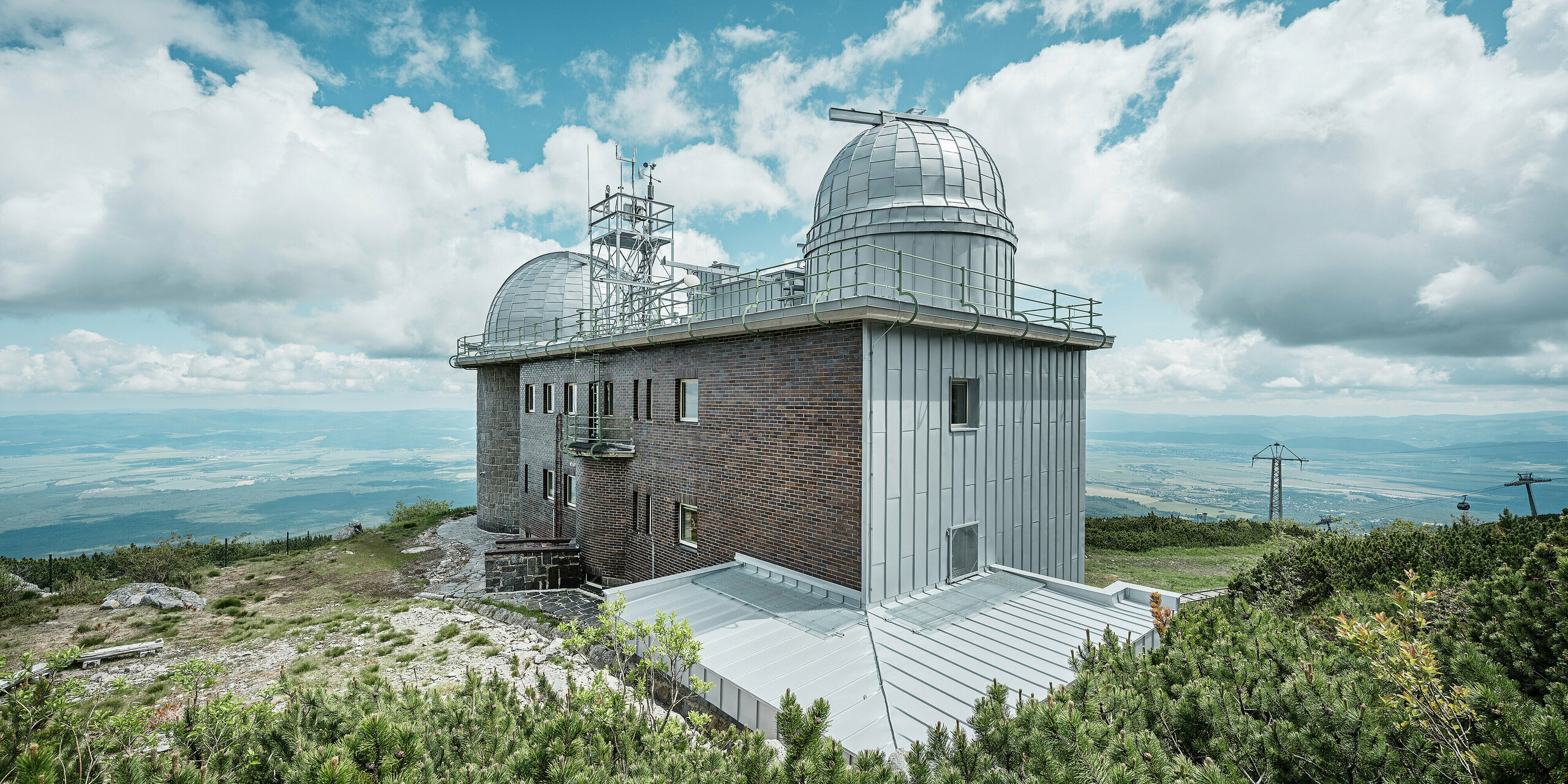 Das Observatorium von Skalnaté Pleso, eingebettet in die majestätische Landschaft der Slowakei, gekrönt mit dem PREFALZ Dachsystem in Silbermetallic. Das denkmalgeschützte Gebäude, eine Symbiose aus solider Ziegelbauweise und modernen Aluminiumelementen, steht stolz auf einem Gipfel in der Hohen Tatra. Die PREFALZ Fassade mit Stehfalz an Dach und Fassade fügt sich nahtlos in die Silhouette ein und bietet dank ihrer Beschaffenheit dauerhaften Schutz vor Witterungseinflüssen und unterstreicht den Blick auf das weite Tal und die sanften Hügel im Hintergrund.