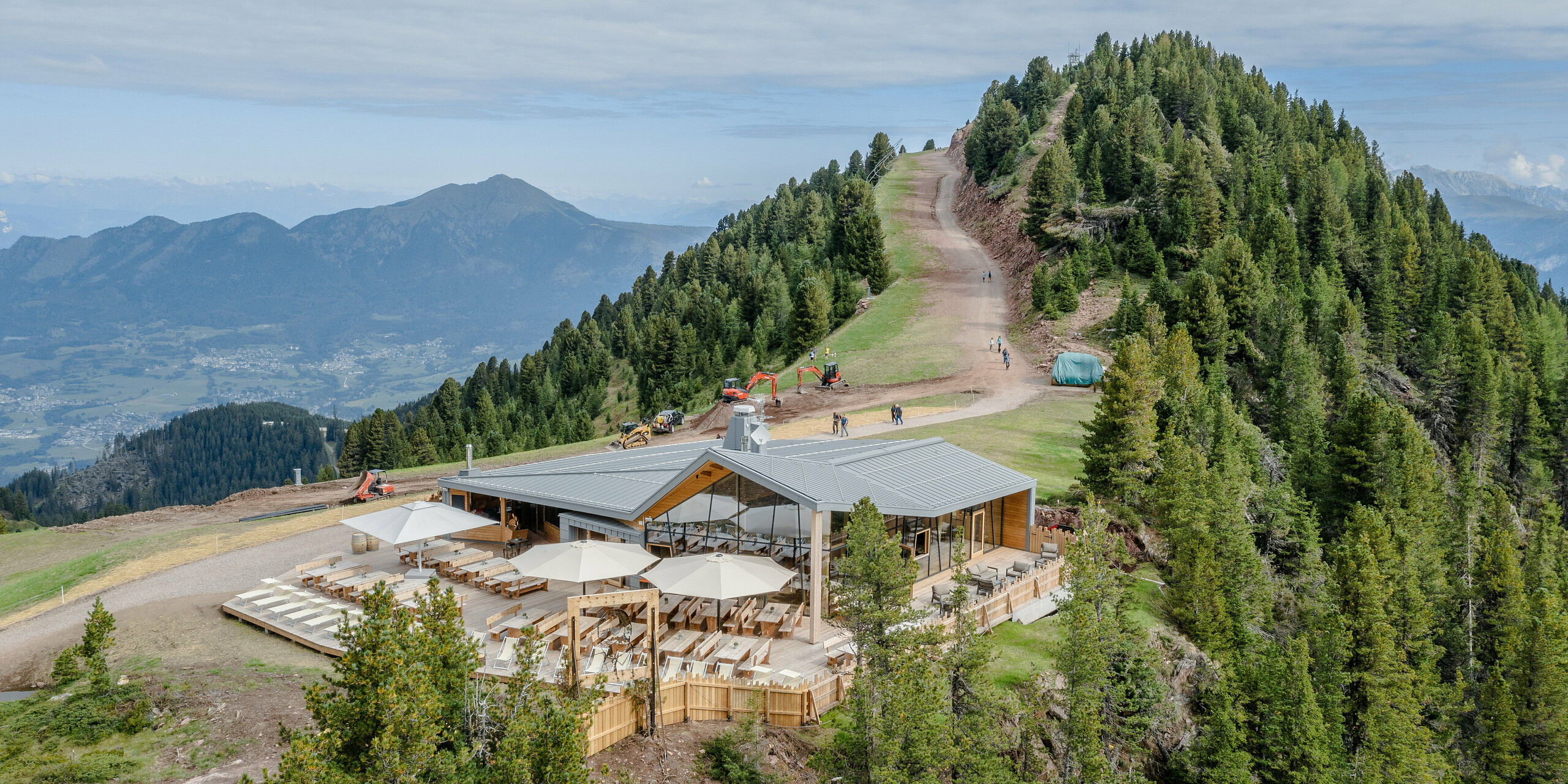 Eindrucksvolle Ansicht des Ristorante Busabelle, gelegen auf einem Bergkamm mit atemberaubendem Panoramablick auf die umliegenden Berge und Täler. Das Restaurant zeigt eine Kombination aus moderner PREFALZ Metallfassade in Hellgrau und natürlichen Holzelementen, eingebettet in eine alpine Landschaft. Die Terrasse des Restaurants ist mit Sonnenschirmen und Holzbänken ausgestattet, die eine einladende Umgebung für Gäste bieten, um die spektakuläre Aussicht zu genießen.